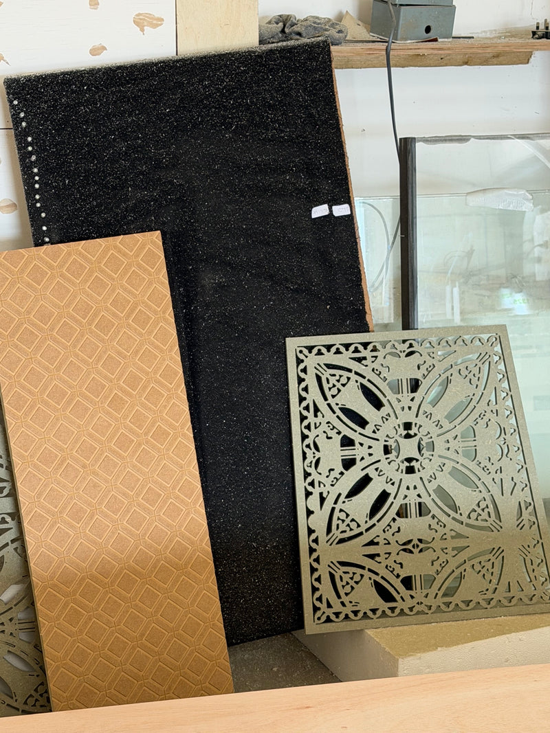 A collection of decorative tiles and panels displayed on a shelf, including a black textured panel, a tan geometric patterned tile, and a sage green cnc-cut mdf panel with an intricate butterfly design.