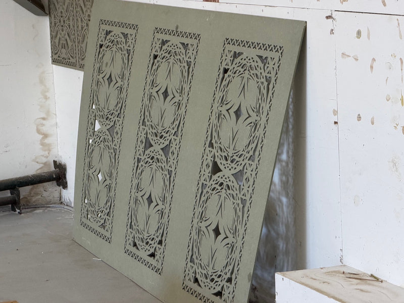 A large decorative panel with three vertical floral cutout sections in a muted sage green color, leaning against a white wall in an unfinished room.