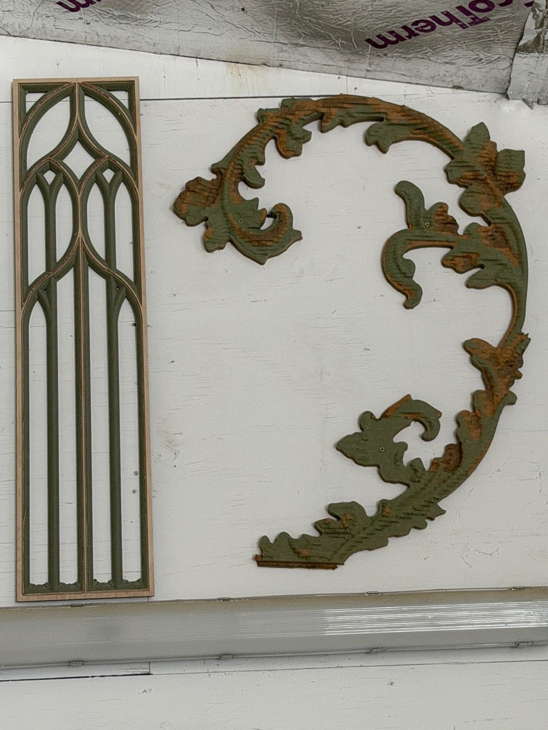 Two vintage decorative wood trim pieces displayed side-by-side on a white wall. The left piece is a tall, narrow rectangular panel with a Gothic arch design, and the right piece is a large, curved leafy acanthus scroll ornament.