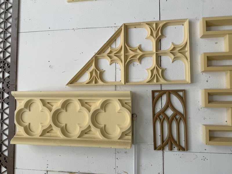 Several decorative architectural molding pieces with Gothic and quatrefoil patterns displayed against a white paneled wall. The pieces are shown in light beige and medium brown tones.