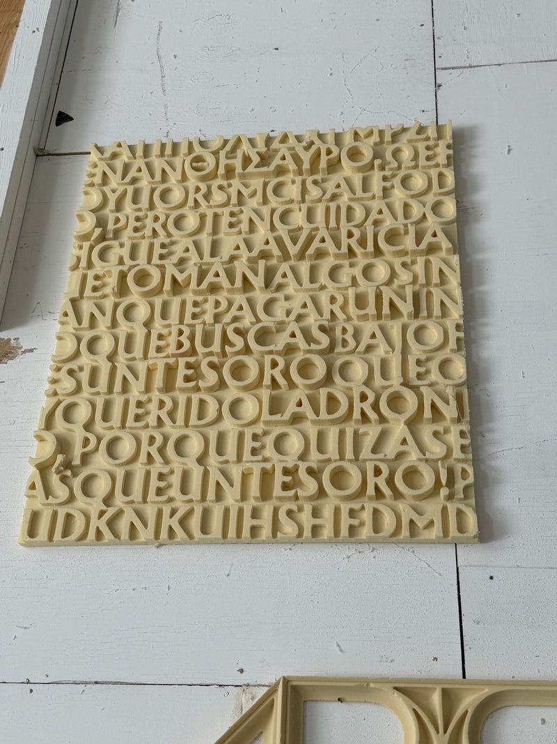 A square beige plastic alphabet board or letterpress plate with rows of raised Spanish letters, photographed against a white wooden background.
