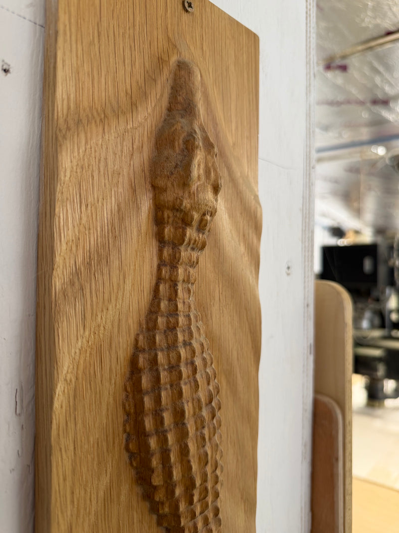 A close-up of a carved wooden wall plaque featuring a stylized crocodile design, mounted on a white wall with a blurred interior space visible in the background.
