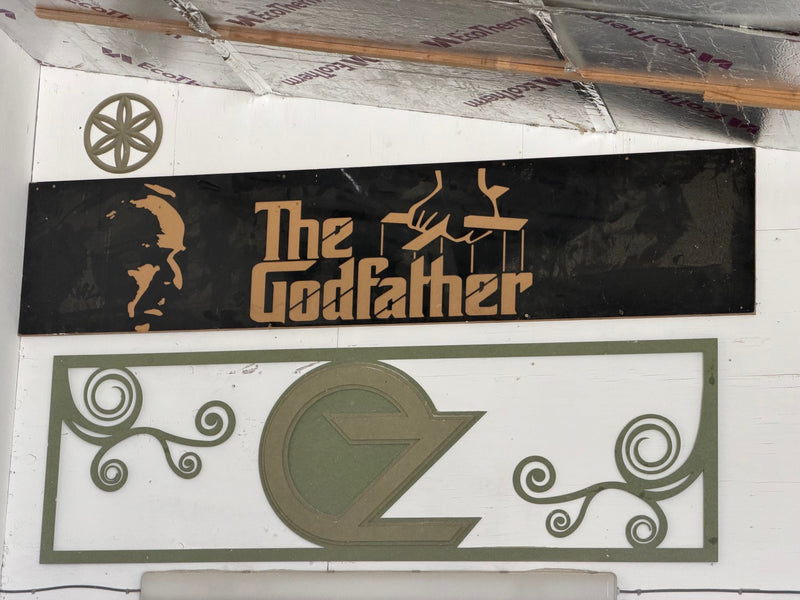 A black rectangular carved panel featuring the 'The Godfather' logo in gold, mounted on a white wall above a larger olive green decorative sign with the stylized 'OZ' logo.