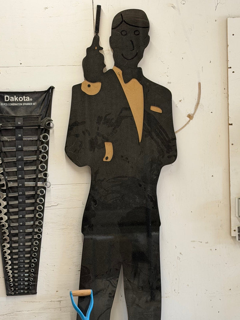 A black silhouette cutout of a man in a suit holding a gun, displayed against a white workshop wall. A blue-handled shovel is at the bottom, and a rack of combination spanners and a white electrical panel are visible on the left and right.